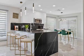 Una cocina moderna con una isla central, taburetes de bar, una zona de comedor con sillas verdes y un amplio ventanal con persianas.