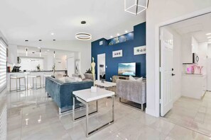 A modern living room with a blue sofa, a white coffee table, and a grey armchair. There is a kitchen area with white cabinets and a dining table. - Luxury Home/Heated Pool/TPA Airport/Disney (Tampa)
