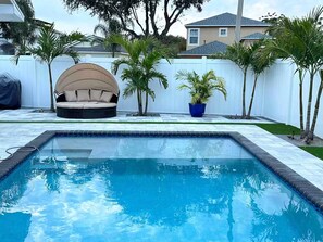 Ein rechteckiger Swimmingpool mit weiß gefliestem Rand, umgeben von einem weißen Zaun, Palmen und einem überdachten Sitzbereich im Freien.