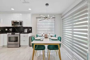 Une cuisine moderne dotée d’appareils électroménagers intégrés, un coin repas avec une table pour deux personnes, et de grandes fenêtres équipées de stores.