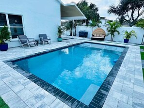 Une piscine rectangulaire dont les bords sont recouverts de carreaux, entourée d’un patio aménagé avec des chaises longues et des plantes en pot.