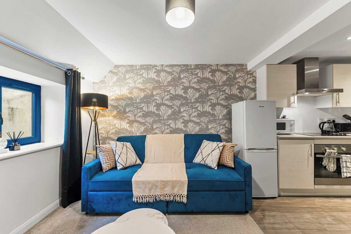 A compact living space with a blue sofa, a floor lamp, a kitchenette, and a window with curtains.