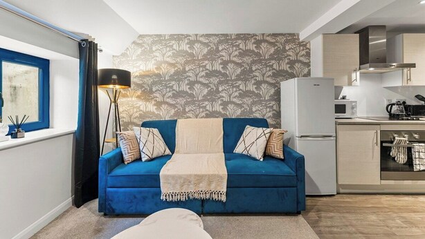 Un espace de vie compact comprenant un canapé bleu, une lampe sur pied, un coin cuisine et une fenêtre avec des rideaux.