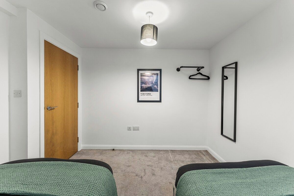 Quarto com porta de madeira, um quadro na parede, um cabide preto, um espelho e duas camas verdes.