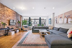 A modern living room with a brick wall, a large window, and a grey sectional sofa. - Spacious 2-bdrm apartment in vibrant NY's East Village Available Aug 12- Sept 30 (New York)