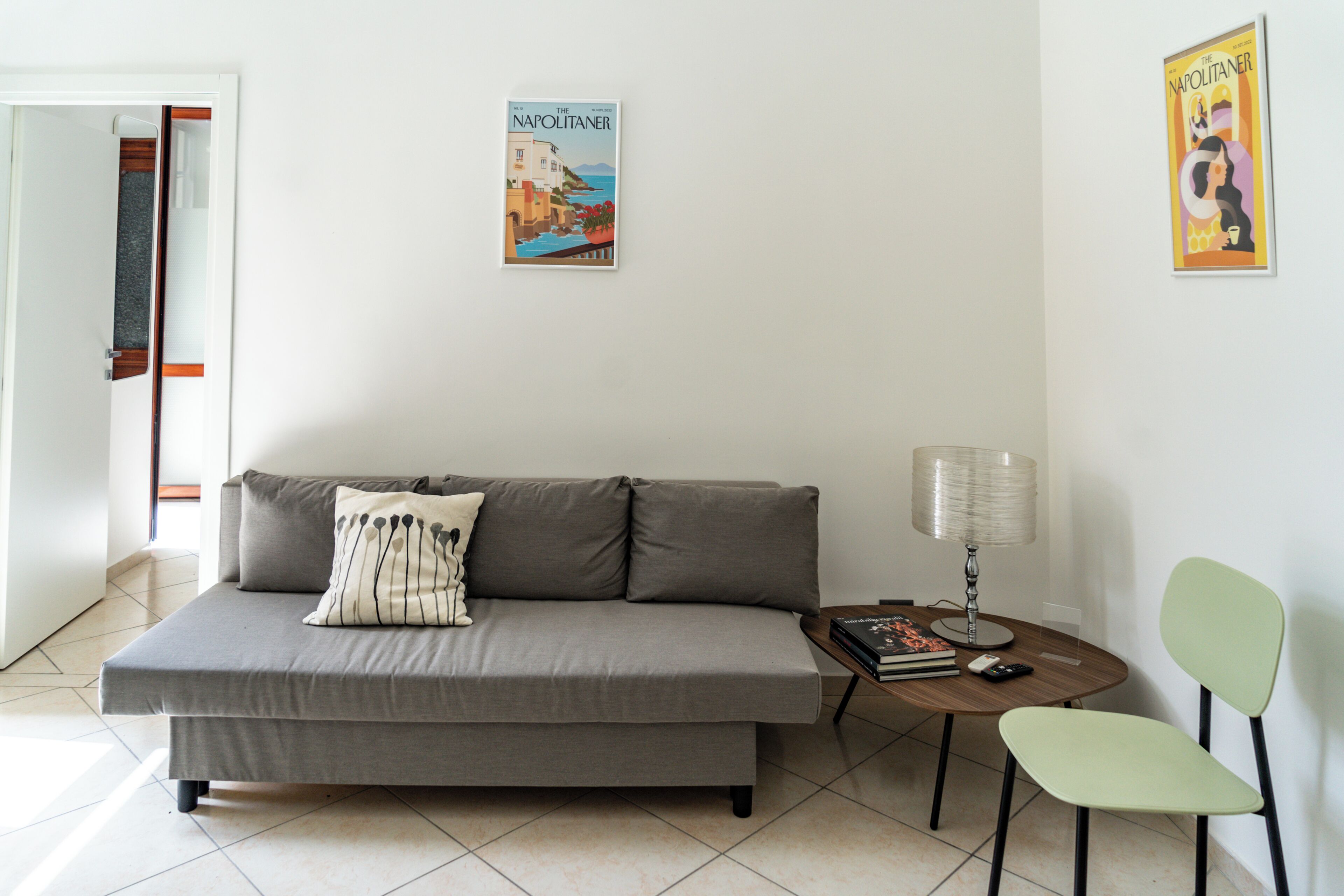 Una sala de estar moderna con un sofá gris, una mesita redonda con una lámpara, una silla y dos carteles enmarcados en la pared.