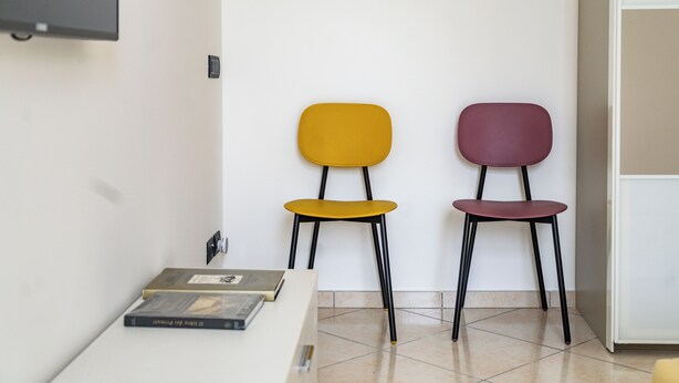 Twee stoelen in verschillende kleuren, een witte tafel met een boek, en een witte muur met een lichtschakelaar.