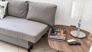 Een moderne woonkamer met een grijze bank, een klein rond houten tafeltje met een lamp, boeken en een afstandsbediening.