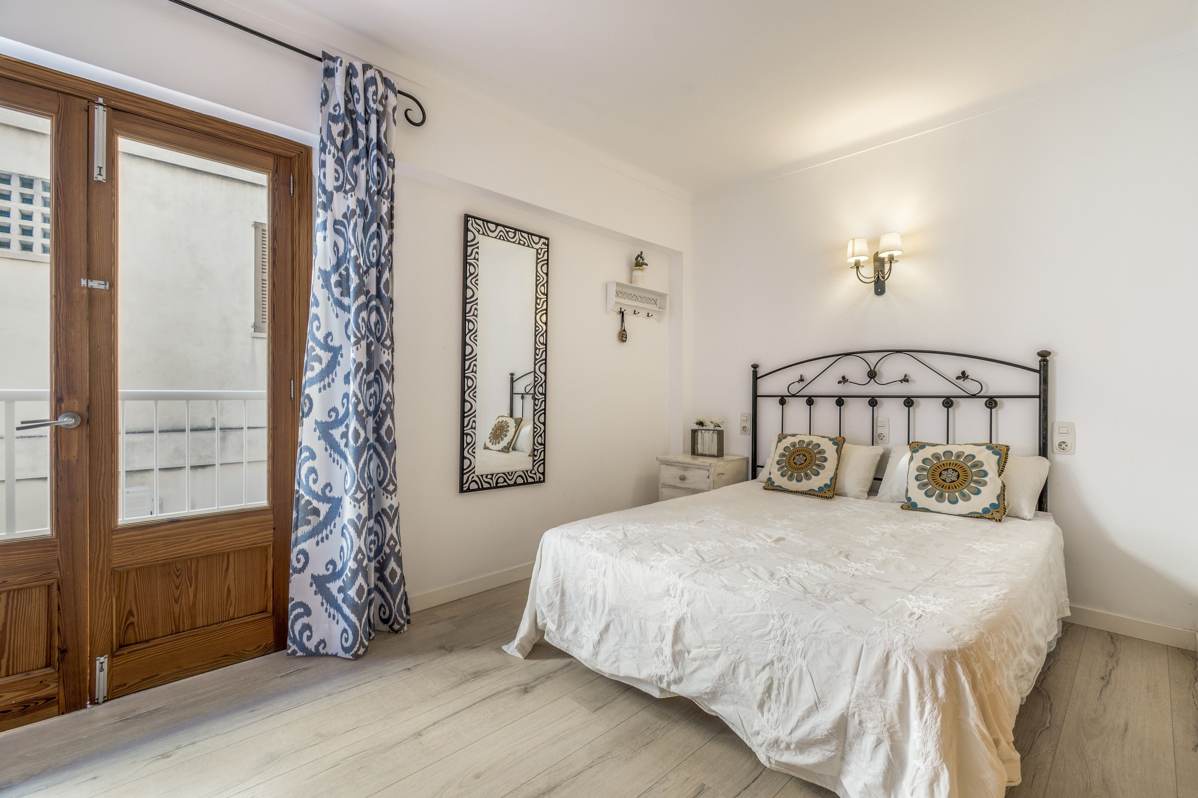 Un dormitorio con puerta de madera, cortina estampada, cama con almohadones decorativos, mesita de noche y espejo montado en la pared.