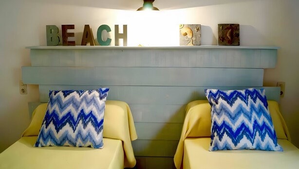 Ein Zimmer mit zwei Betten, jedes mit einem blau-weißen Zickzackmuster-Kissen, einem Regal darüber mit dem Wort 'BEACH' in Buchstaben und einem kleinen dekorativen Gegenstand auf dem Regal.
