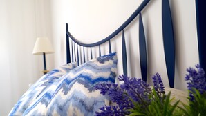 Ein Bett mit einem blau-weißen Kissen, einer beigen gesteppten Decke und einer Vase mit lila Blumen auf einem Nachttisch.