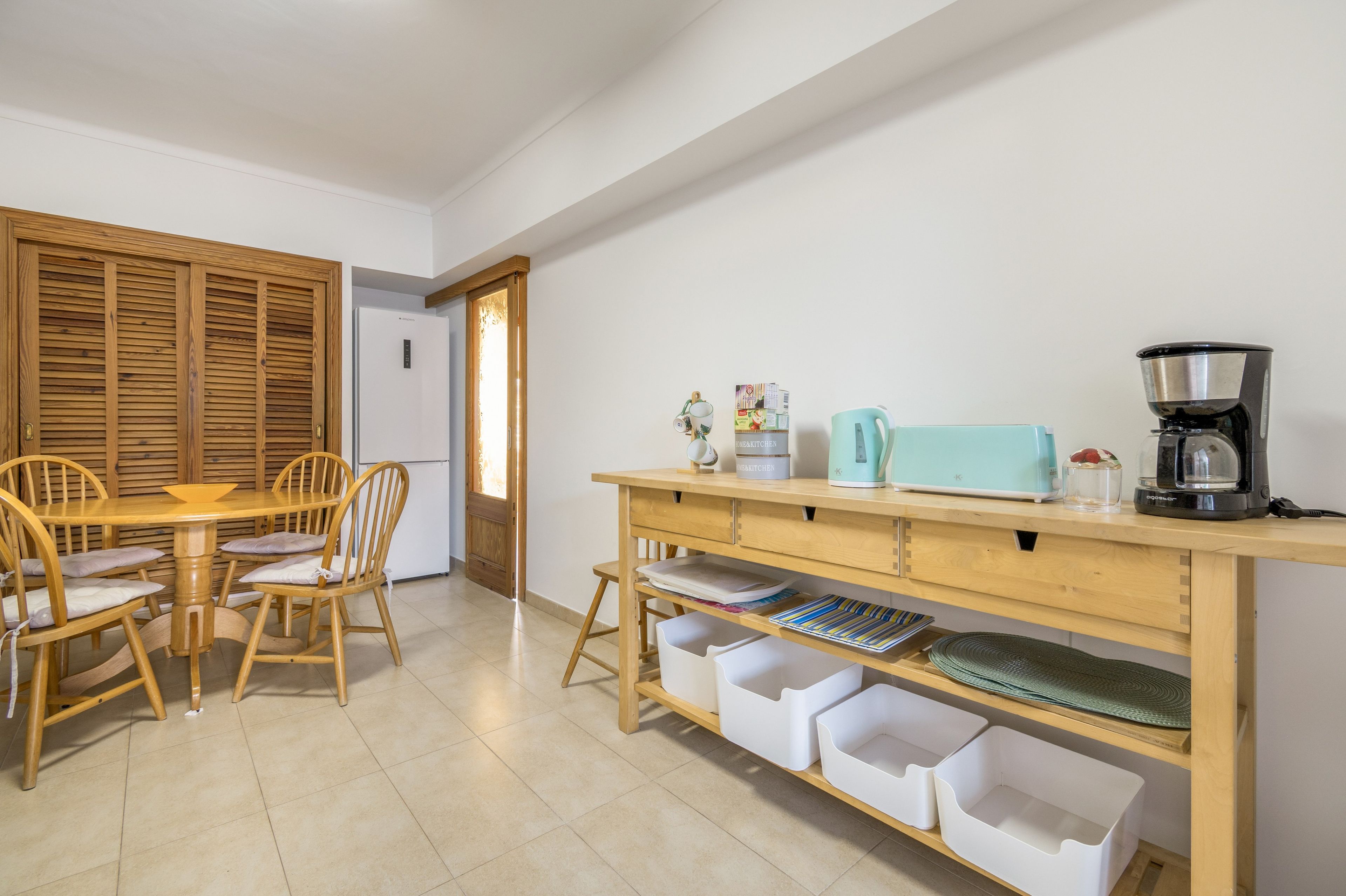 Un comedor con una mesa y sillas de madera, una encimera de cocina con electrodomésticos y contenedores de almacenamiento.