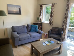 A living room with a sofa, a chair, a coffee table, a lamp, a bookshelf, and a window with curtains. - 6 Riverfalls Clifden (Clifden)