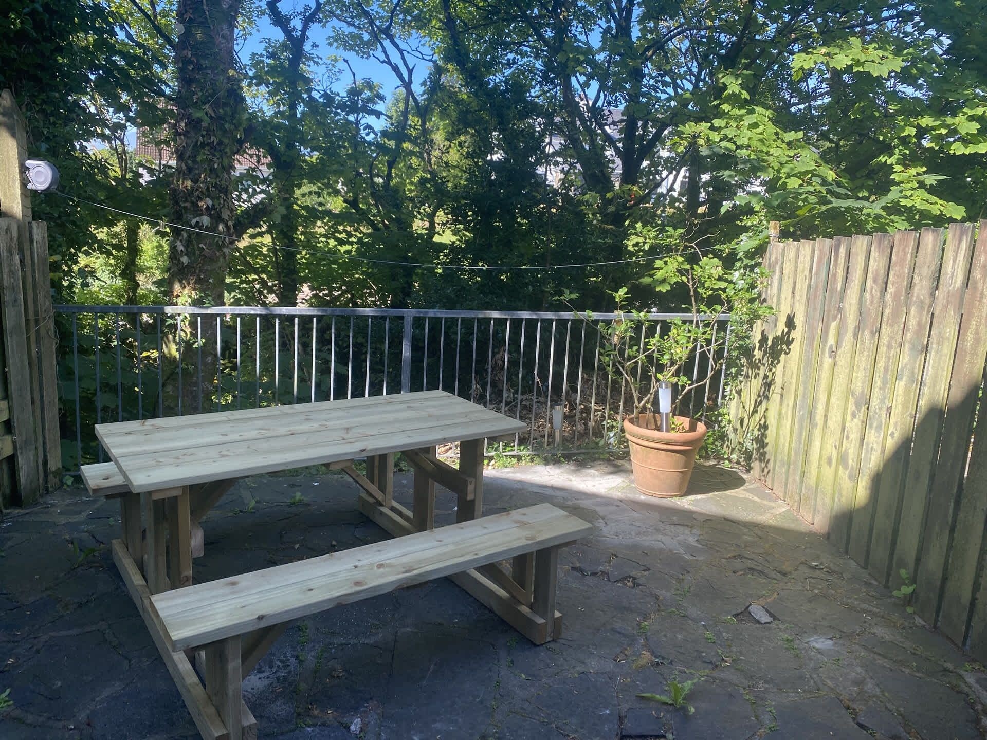 Een houten picknicktafel met bankjes in een tuin, met een metalen hek en een bloempot.