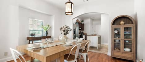 A dining area with a wooden table, white chairs, a vase with flowers, and a kitchen in the background.