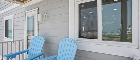 En veranda med to blå Adirondack-stole, et hvidt gelænder og et vindue med udsigt til en bygning.