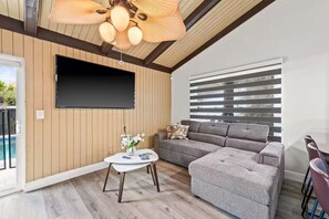 A modern living room with a grey sectional sofa, a flat-screen TV mounted on the wall, a ceiling fan, and a pool visible through the glass door. - Fancy Tampa Retreat/Disney/Heated Pool/Clearwater (Tampa)