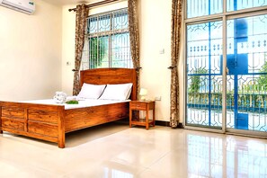 A bedroom with a wooden bed, a bedside table, and a view of a blue gate through a window. - Beachfront Apartment, Terrace Onto Sand Uroa Zanzibar (Uroa)