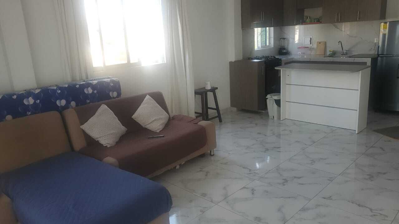 A living room with a sofa, a small table, and a kitchen area with a refrigerator and a sink.