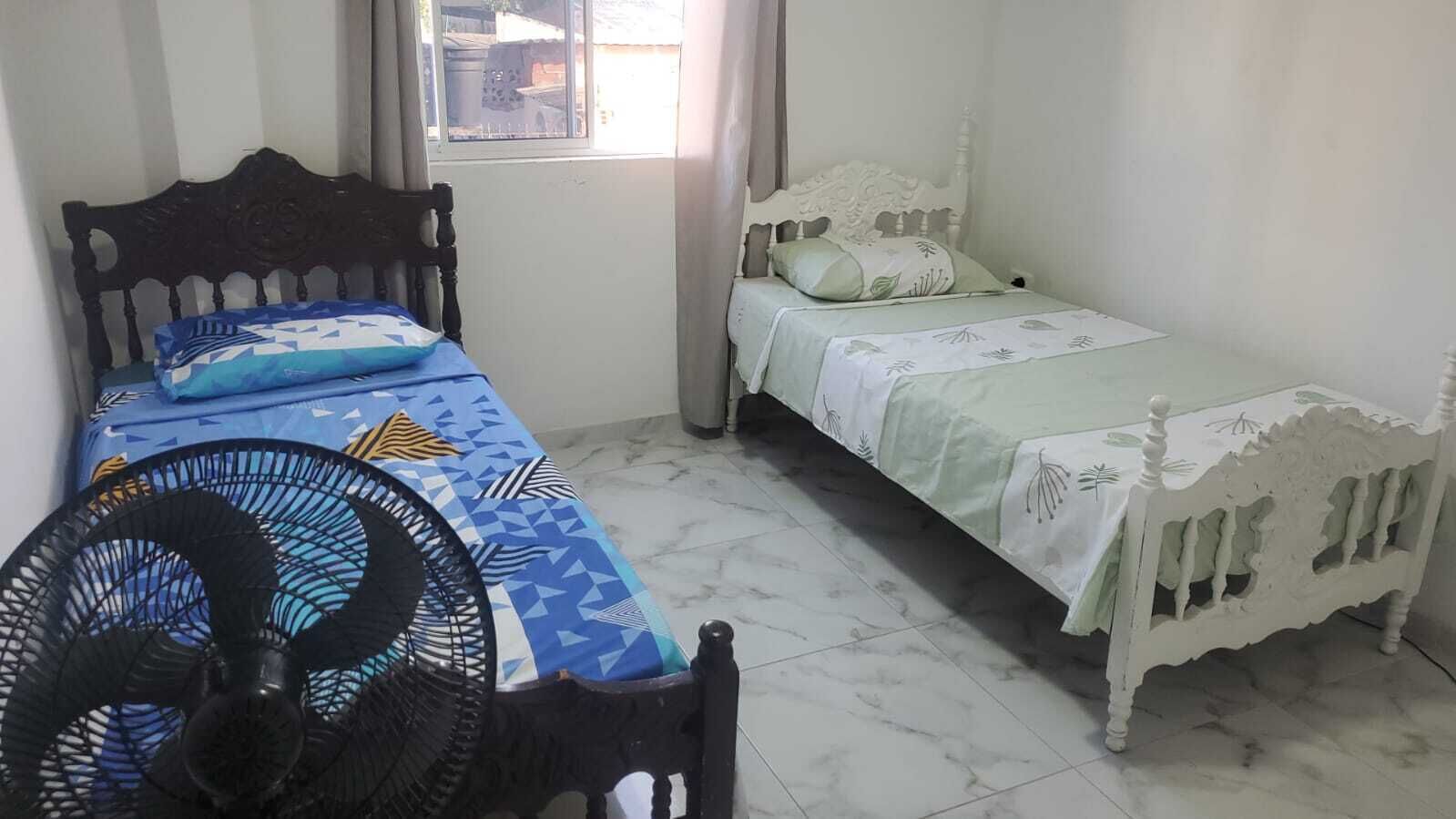 Ein Zimmer mit zwei Betten, eines mit einer gemusterten Tagesdecke und das andere mit einer schlichten. Im Vordergrund steht ein Standventilator.