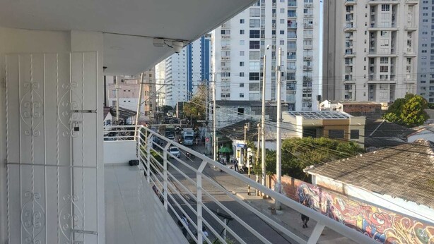 Un balcon doté d’une rambarde en métal, donnant sur une rue animée bordée d’immeubles et traversée par des véhicules.