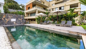 Piscina con sillas de descanso y sombrillas, rodeada de un muro de piedra y vegetación.