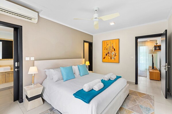A hotel room with a bed, bedside tables, a ceiling fan, a painting, and a door leading to another room. - 17 Ocean Legend (Tamarin)