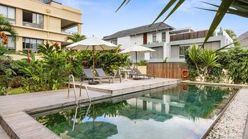 A swimming pool with lounge chairs and umbrellas, surrounded by lush greenery and modern buildings.
