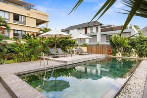 A swimming pool with lounge chairs and umbrellas, surrounded by lush greenery and modern buildings. - 17 Ocean Legend (Tamarin)