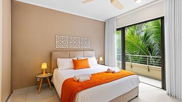 A hotel room with a bed, nightstands, a ceiling fan, and a view of greenery through large windows.