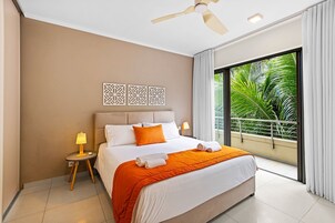 Habitación de hotel con una cama, mesitas de noche, ventilador de techo y vistas a zonas verdes a través de grandes ventanales.