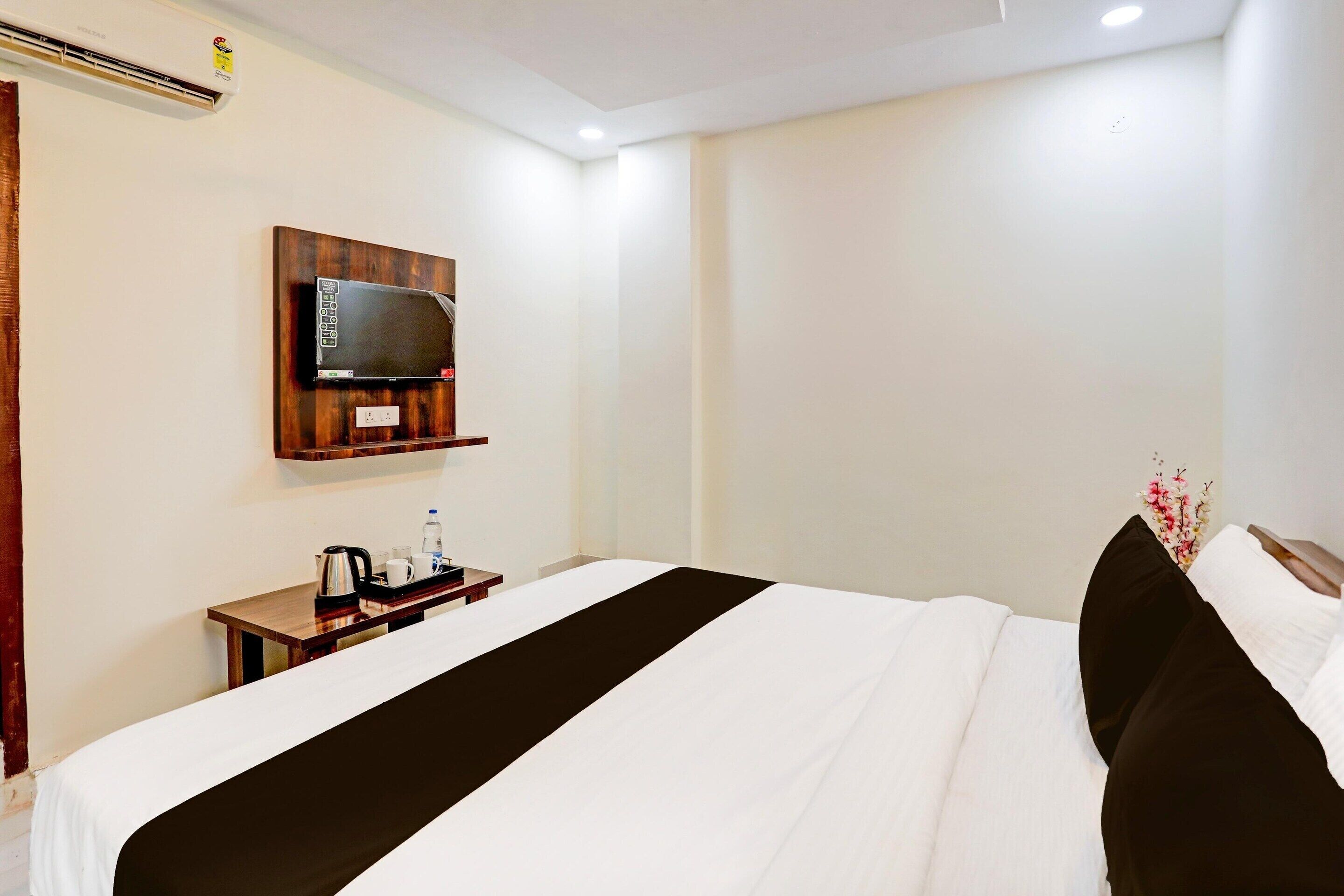 Habitación de hotel con cama, mesita de noche con tetera y tazas, televisor de pantalla plana montado en la pared y un pequeño estante de madera.