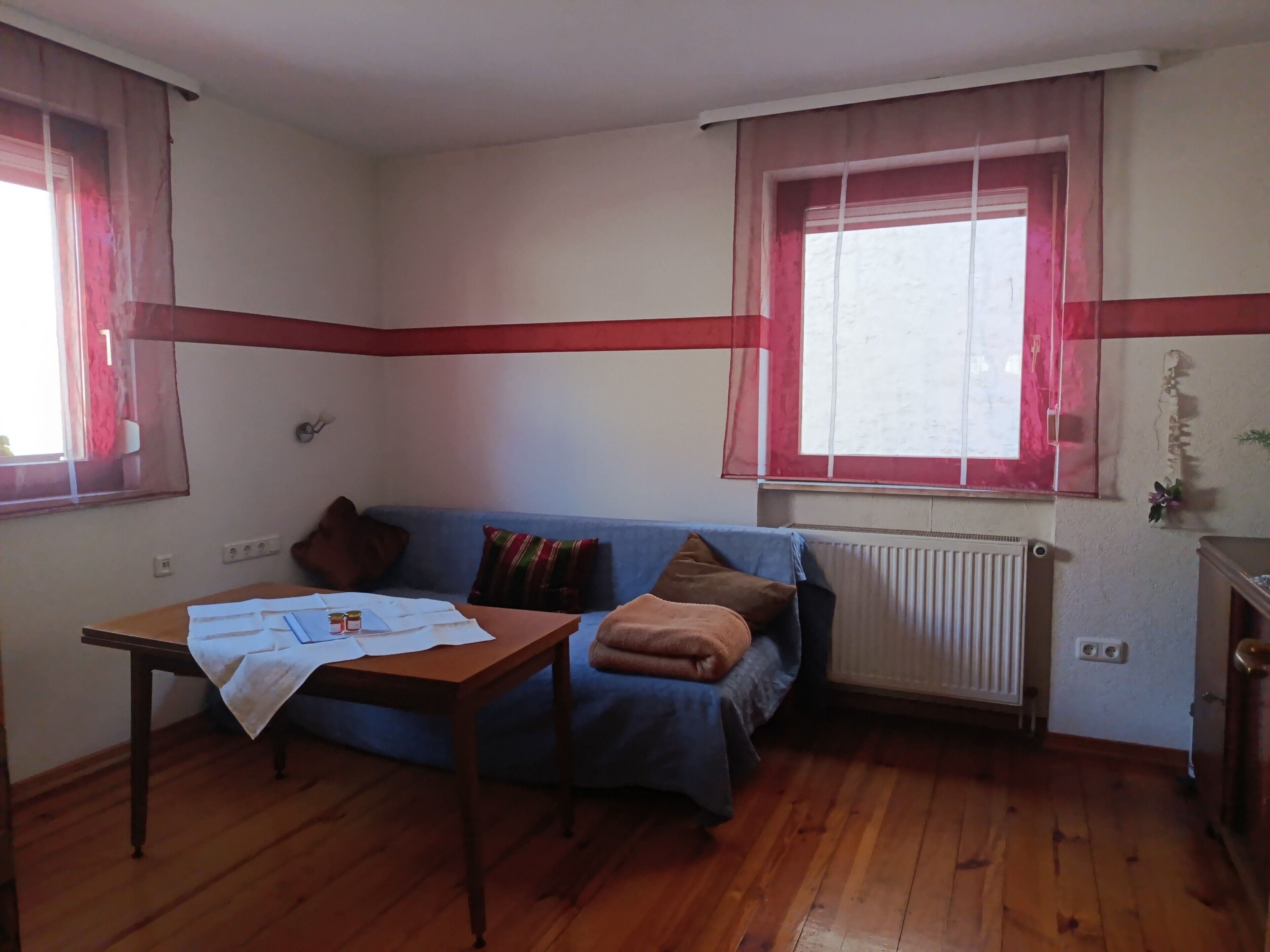 Una stanza con pavimento in legno, un divano blu con cuscini, un tavolo con documenti e una finestra con tende.