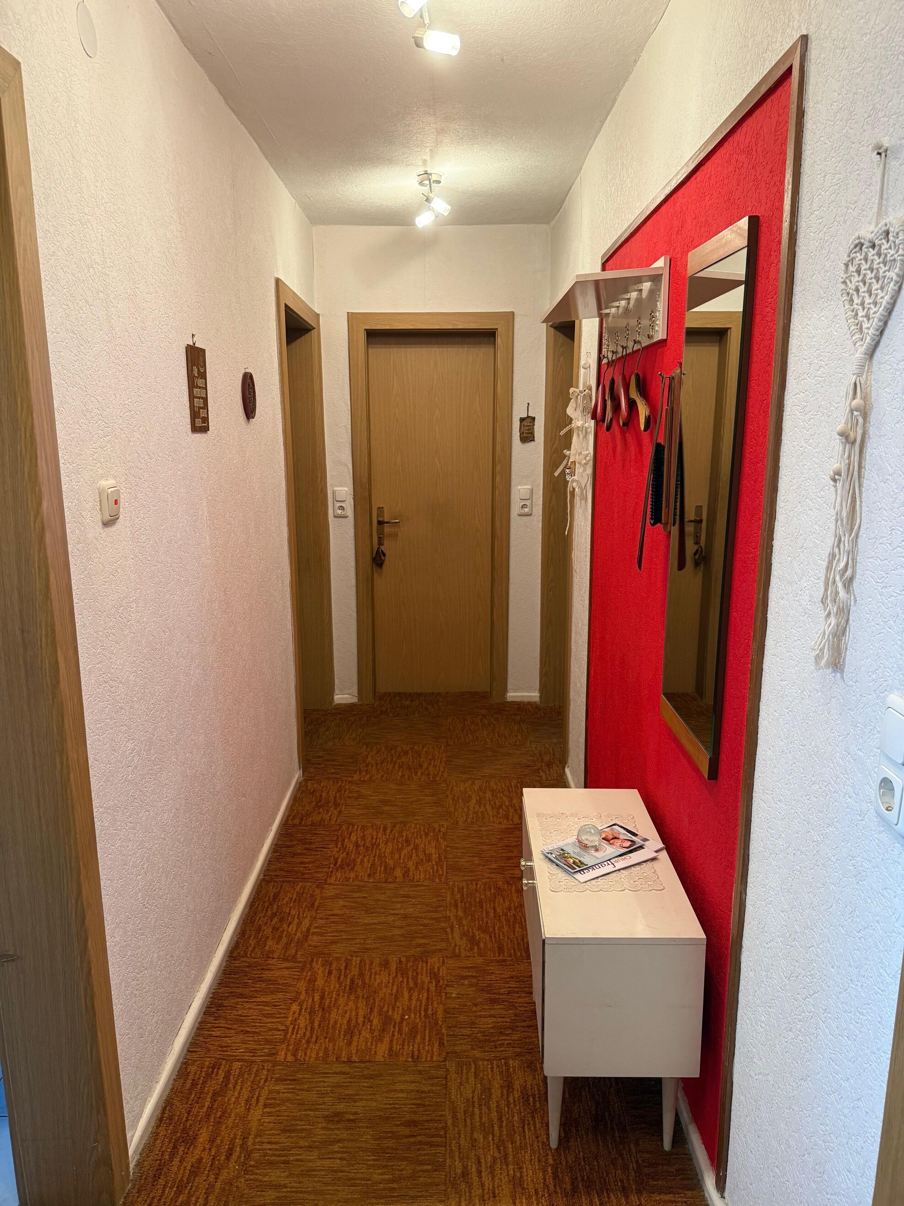 A hallway with a wooden door at the end, a white bench with magazines, and a red wall with a mirror and hanging items.