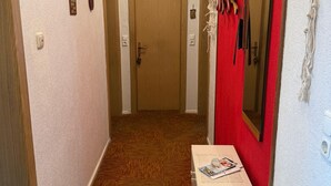 A hallway with a wooden door at the end, a white bench with magazines, and a red wall with a mirror and hanging items.