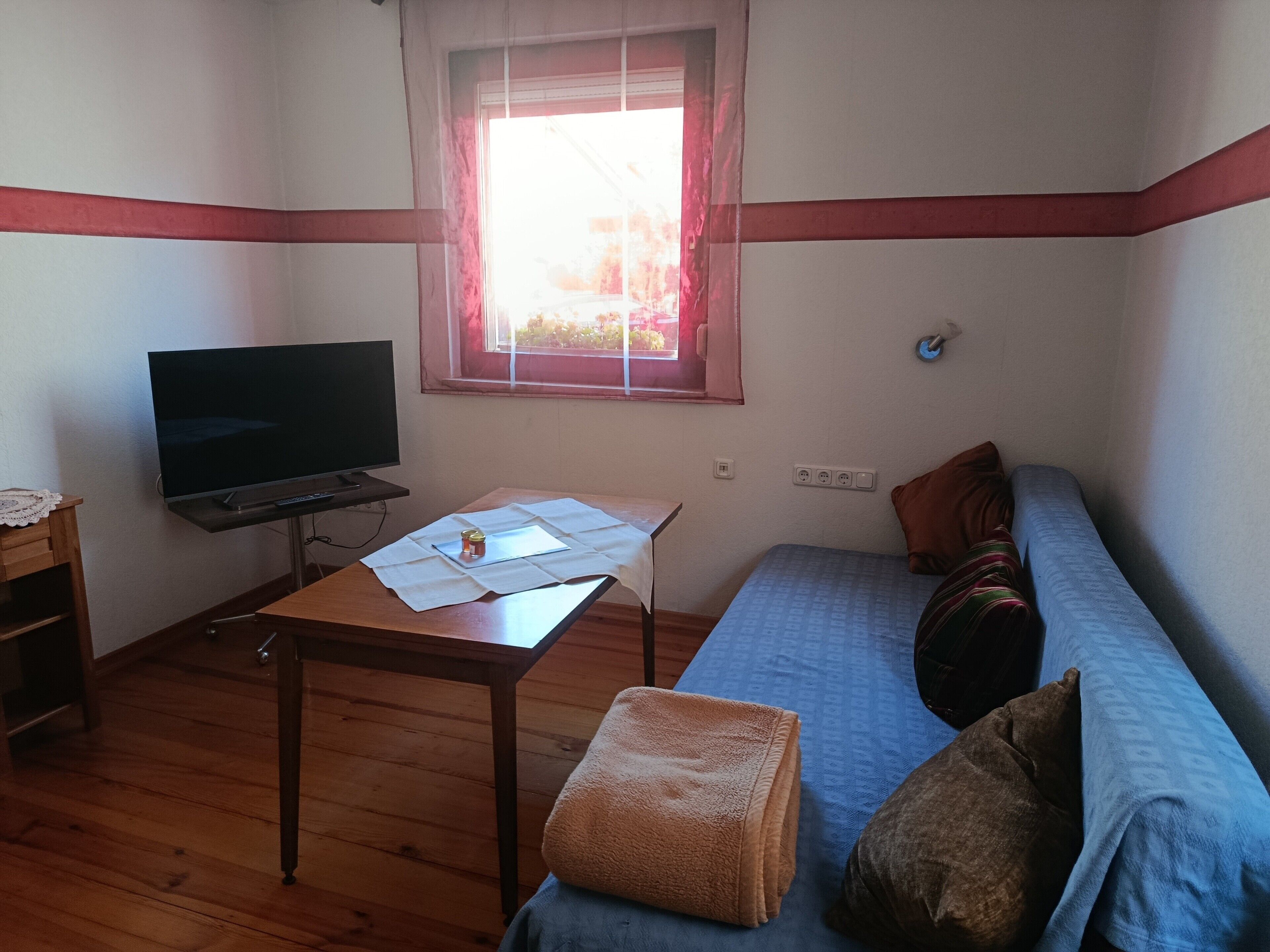 Een kleine, ingerichte kamer met een televisie, een tafel met papieren, een raam met gordijnen, een bank met kussens en een houten vloer.
