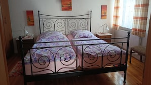 A bedroom with a metal bed frame, a bedside table with a lamp, a window with curtains, and two paintings on the wall.