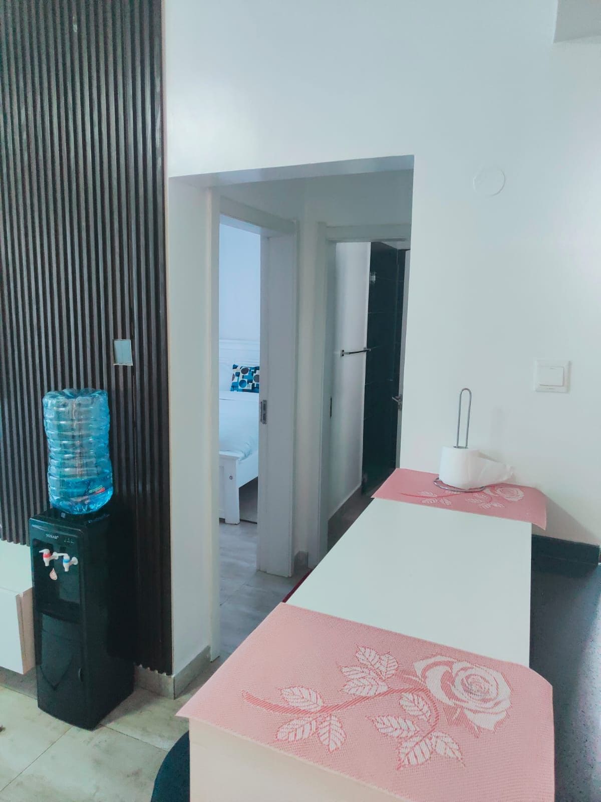 Quarto de hotel com dispensador de água, uma cama, mesa de cabeceira com abajur e vista para o quarto vizinho.