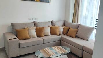 A living room with a sectional sofa, a glass coffee table, a painting on the wall, and a window with curtains.