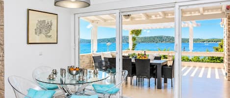Un espace repas moderne avec une table et des chaises en verre, un tableau encadré au mur et une vue sur la mer par de grandes portes vitrées.