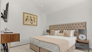 Ein modernes Schlafzimmer mit einer gepolsterten Kopfteil, einem großen Bett, einem Nachttisch mit Lampe, einem Schreibtisch mit Computer und einem gerahmten Kunstwerk an der Wand.