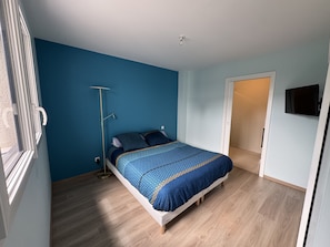Une chambre à coucher avec un lit, une lampe sur pied, un téléviseur fixé au mur et une porte donnant sur une autre pièce.