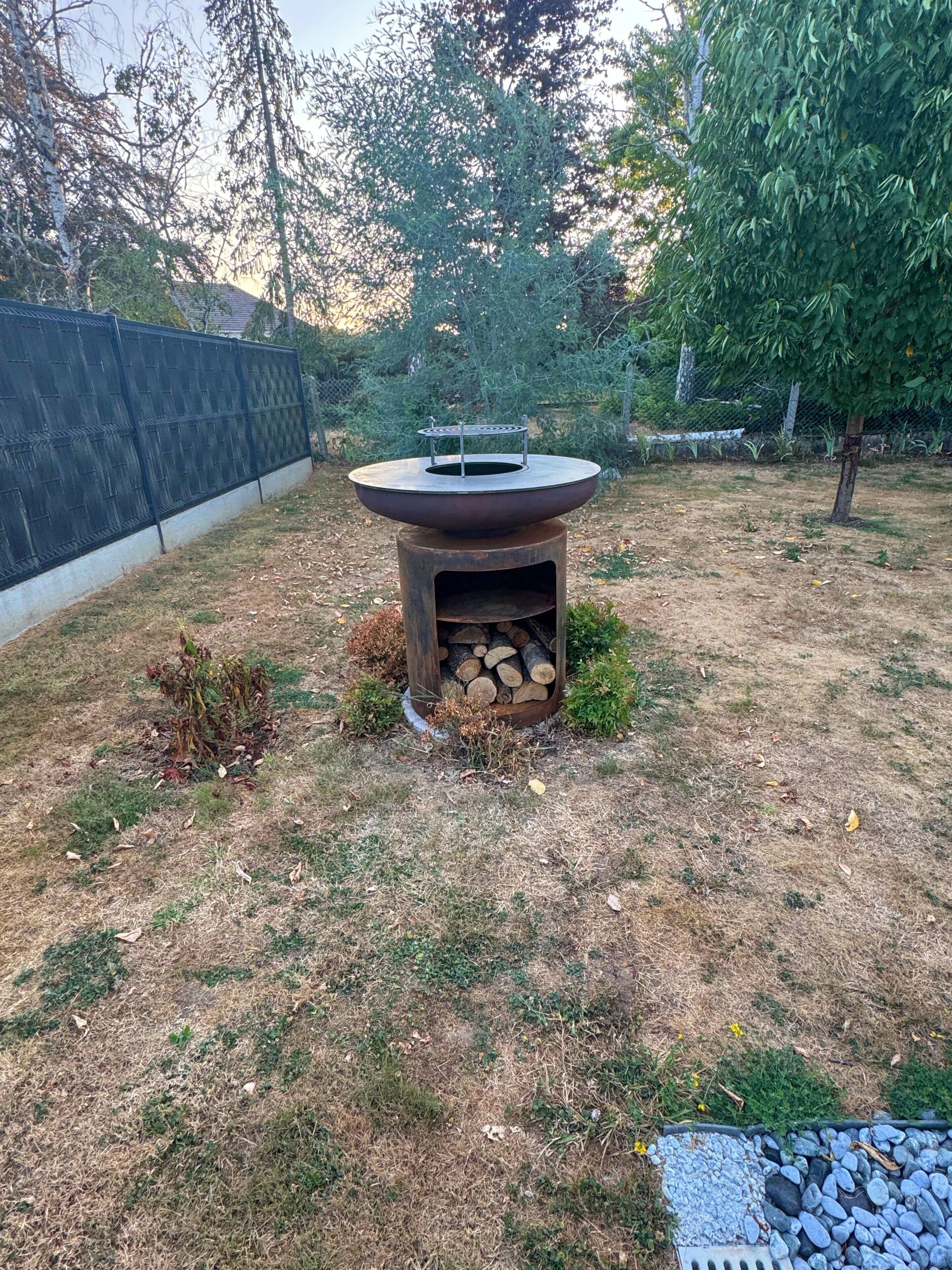 A backyard with a fire pit surrounded by a fence, a small tree, and a grassy area.
