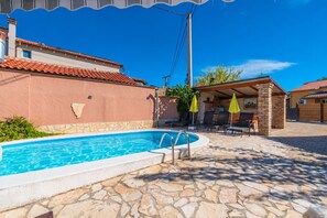 Une piscine avec des carreaux de pierre, un espace extérieur aménagé avec des sièges et un patio couvert.