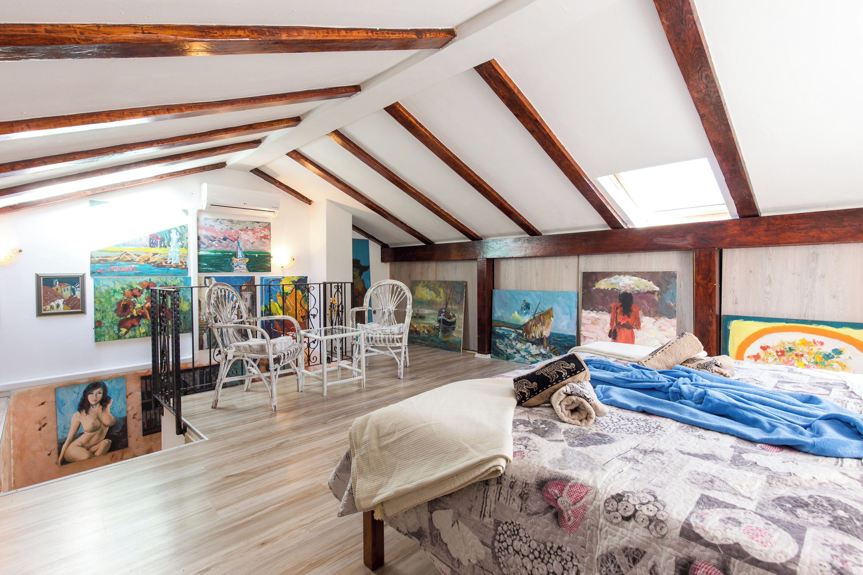 A room with a sloped ceiling, wooden beams, a bed with a patterned bedspread, a chair, and various paintings on the walls.