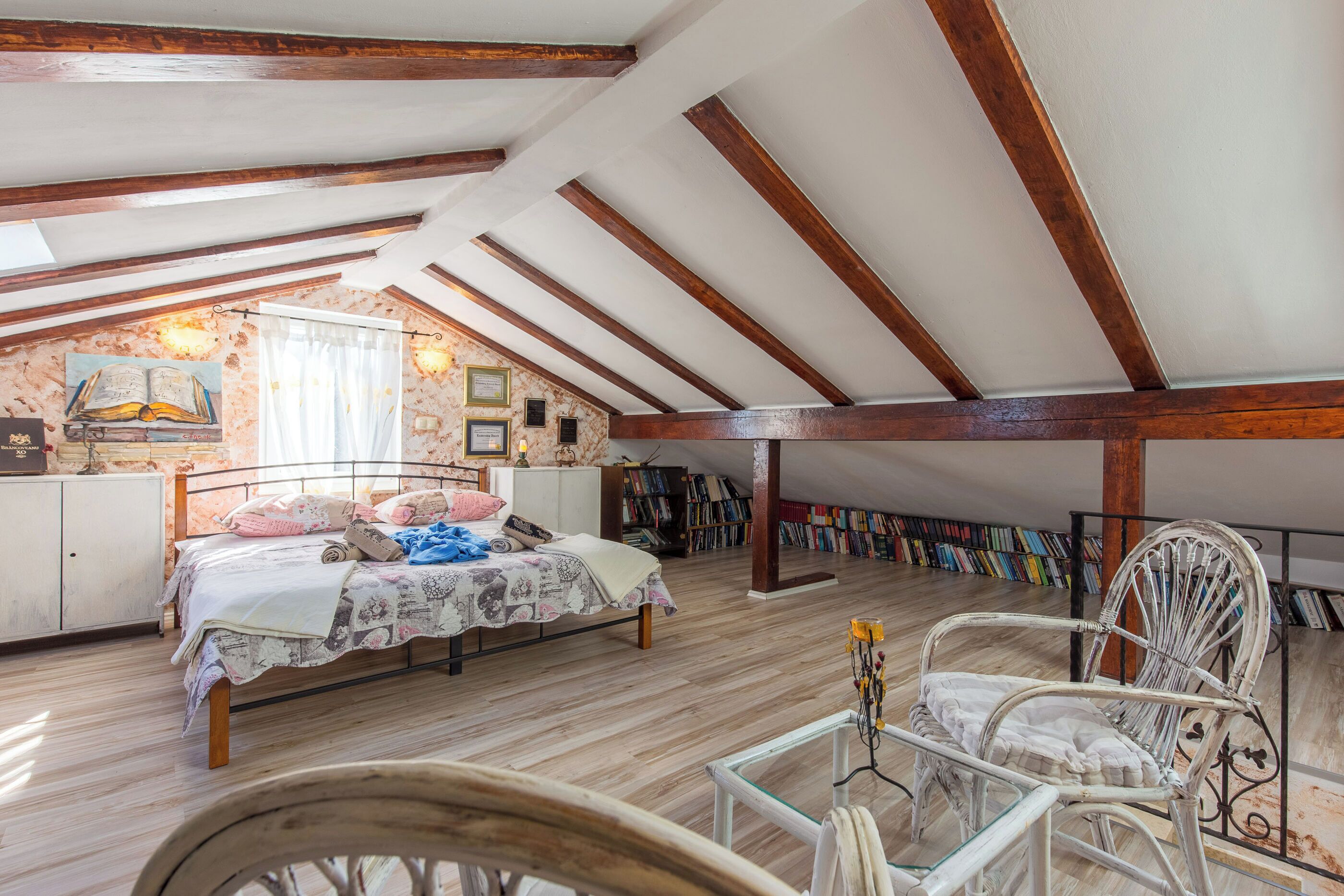 Une chambre avec un plafond en pente, un lit recouvert d’une couvre-lit à motifs floraux, une chaise et une bibliothèque remplie de livres.