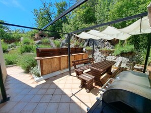 An outdoor patio with a wooden table and benches, a barbecue grill, and a stone wall. - Apartments Emily (Opatija)