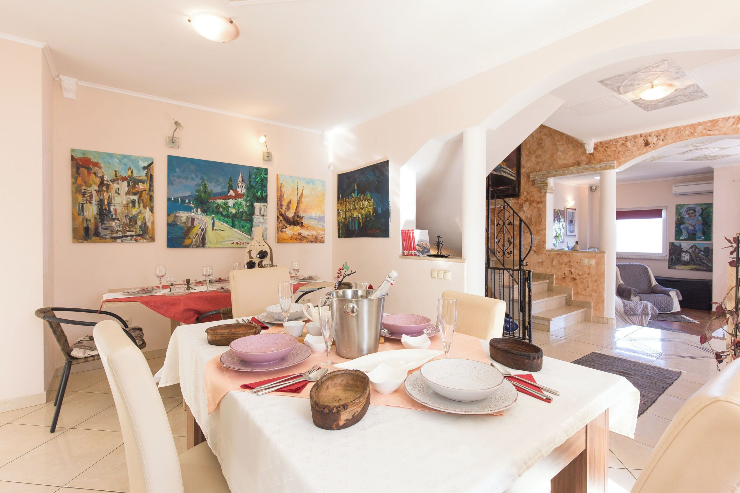 A dining area with a table set for four, a staircase, and a living room with a sofa and a painting.