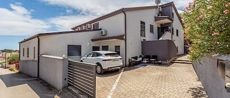 A two-story building with a car parked in the driveway, a gated entrance, and a small outdoor seating area.