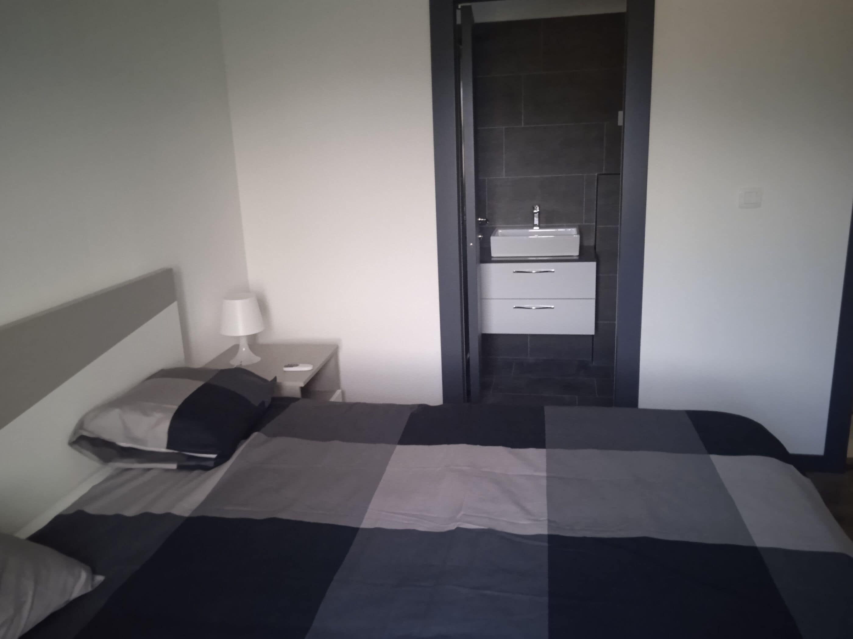 A hotel room with a bed, a nightstand with a lamp, and a bathroom with a sink and drawers.